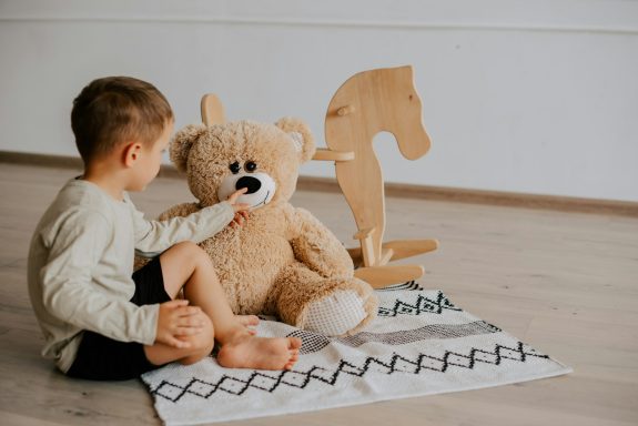 Ein Junge sitzt auf einem Teppich und spielt mit einem großen Teddybär. Im Hintergrund steht ein Holzpferd.