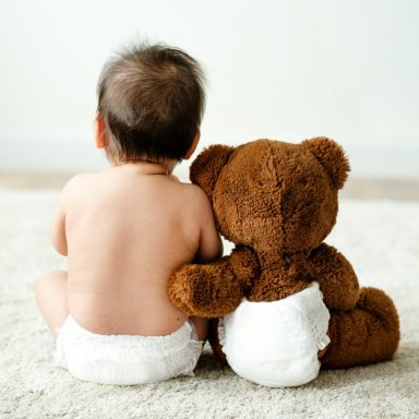 Ein Baby mit nacktem Rücken sitzt neben einem braunen Teddy auf einem Teppich.