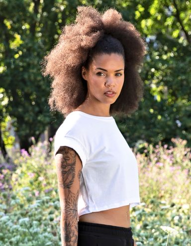 Junge Frau mit lockigem Haar in einem weißen T-Shirt, im Freien vor Pflanzen.