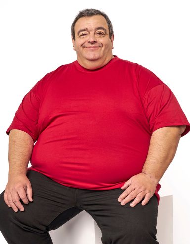 Mann mit breiter Statur, lächelnd, trägt ein rotes T-Shirt und schwarze Hosen.