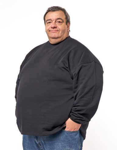 Ein lächelnder Mann in einem schwarzen Sweatshirt und Jeans steht mit einer Hand in der Tasche.